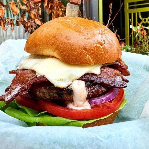 A P'unk Burger with melted cheese, crispy bacon, red onion, tomato, and lettuce, held by a knife.