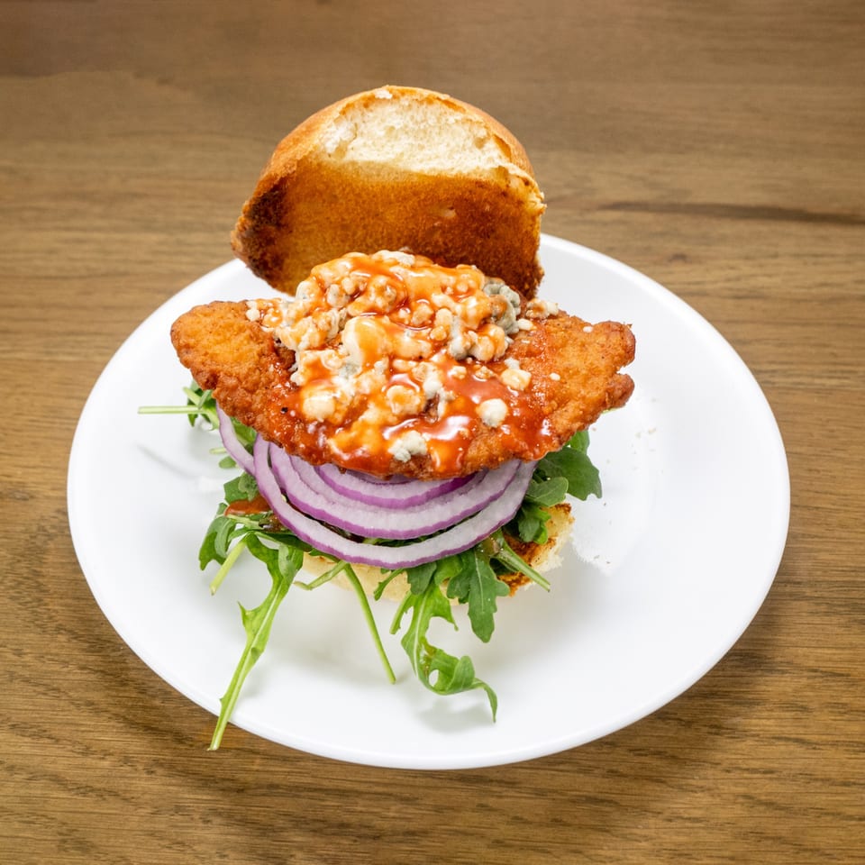 P'unk Burger's buffalo chicken sandwich with blue cheese, red onion, and arugula on a white plate.