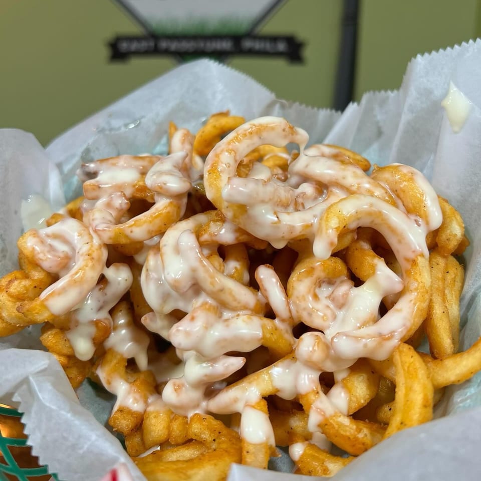 Close-up of P'unk Burger's curly fries, generously topped with creamy white sauce, in a paper-lined basket.
