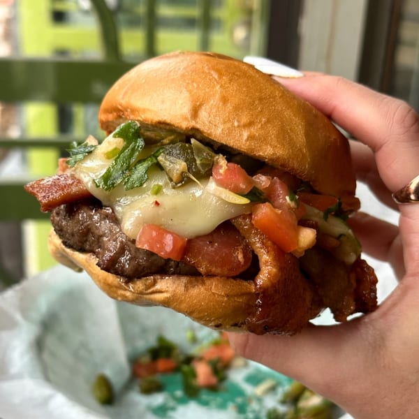 A hand holds a P'unk Burger with melted cheese, bacon, diced tomatoes, and green toppings.